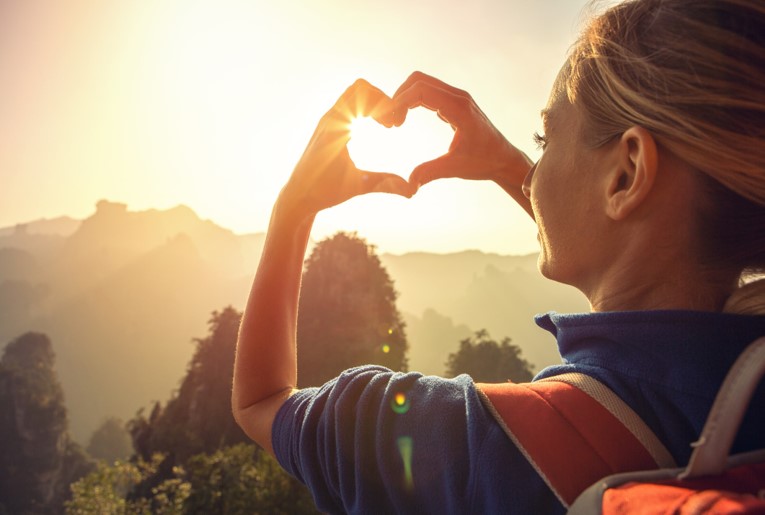 A woman forming a heart shape with her hands around a glowing sunset, symbolizing love, connection, and the beauty of nature.